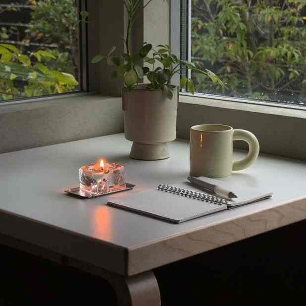 白いテーブルの上に透明なキャンドルとマグカップ、鉢植えが置かれた窓際の落ち着いた雰囲気のある空間