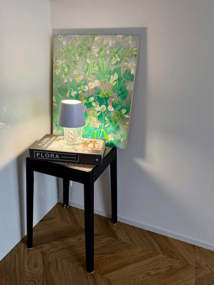 ナチュラルカラーフロアの隅に、キャンドルランプと植物の絵画が置かれた黒いスツールが配された印象的な空間