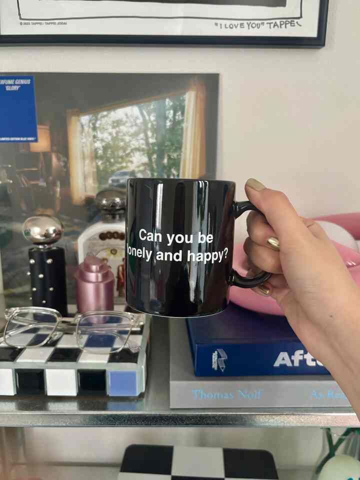 Simple black mug with white text held in a 1LDK bedroom space featuring shelves and assorted accessories