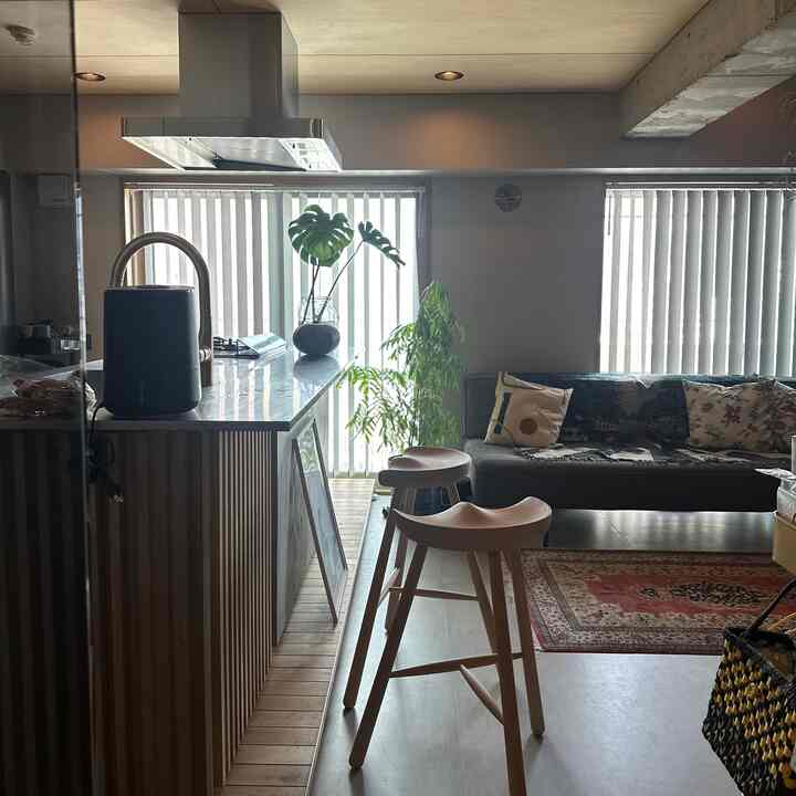 明るい木目調のスツールとグレーのソファ、縦型ホワイトブラインド、緑の植物が配置されたナチュラルモダンなリビングとキッチン空間