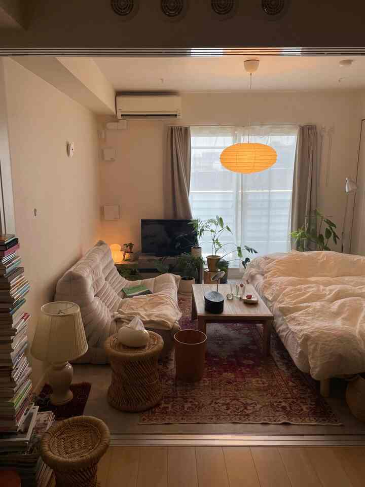 明るいベージュとナチュラルカラーのコンパクトな1LDKリビング、ソファとベッド、植物、自然素材の家具が配置された雰囲気のある空間