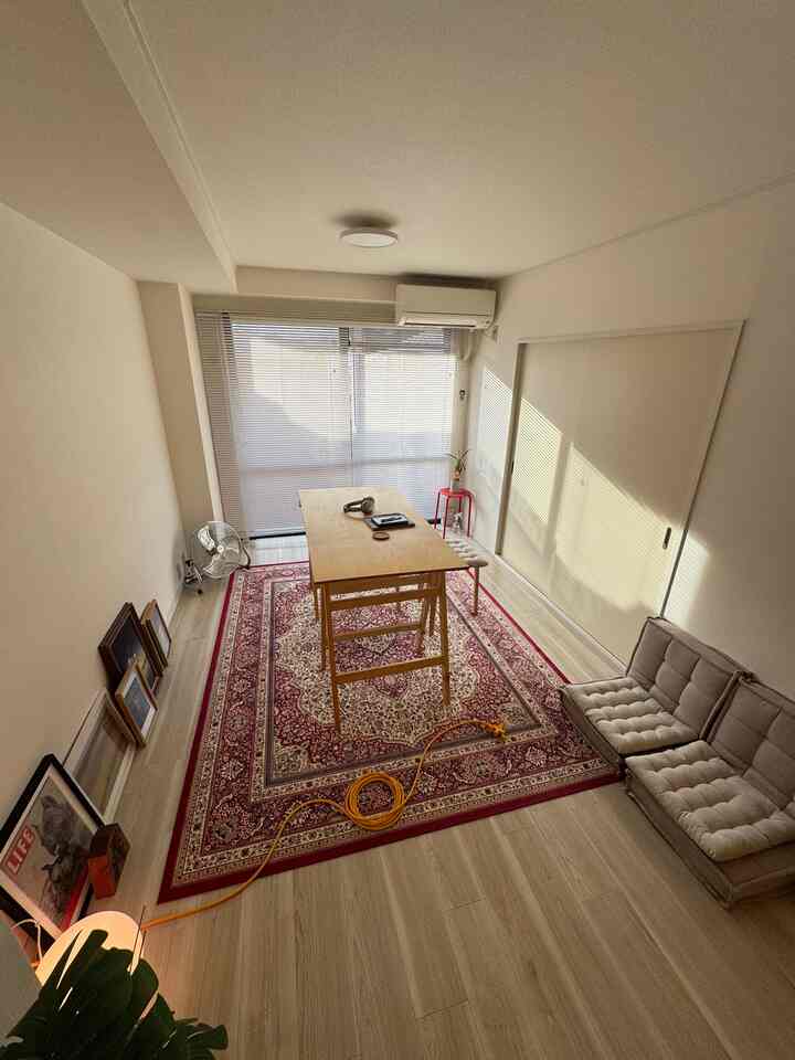 A narrow room with white walls and brown wooden floor, featuring a wooden table and rug at center, small sofa, and warm indirect lighting for a simple single household space