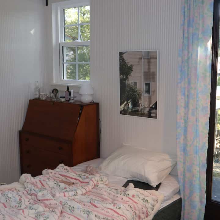 白を基調とした壁と茶色のチェスト、シングルベッド、花柄のカーテンが配置された落ち着いた寝室のインテリア