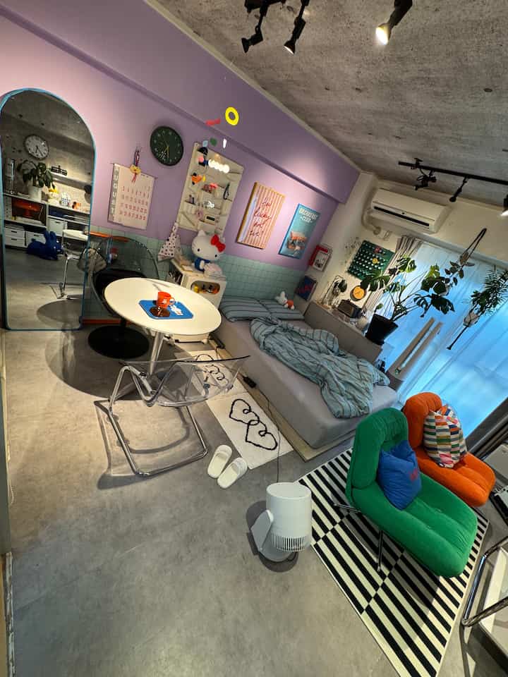 A small living room with purple and gray tones featuring a single bed, round table, and green and orange armchairs in a modern simple setting