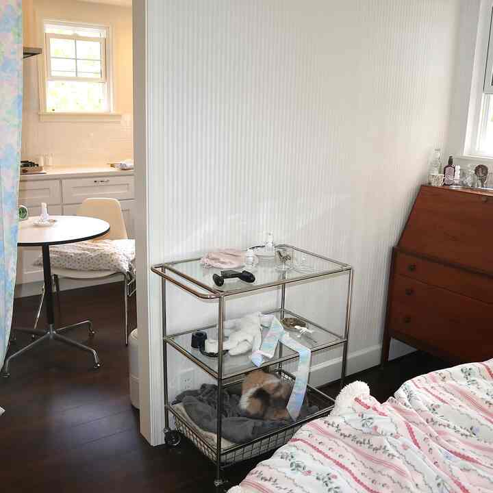 White and brown-toned studio apartment kitchen and bedroom space featuring a cat on a pet bed inside a metal storage cart, creating a cozy atmosphere