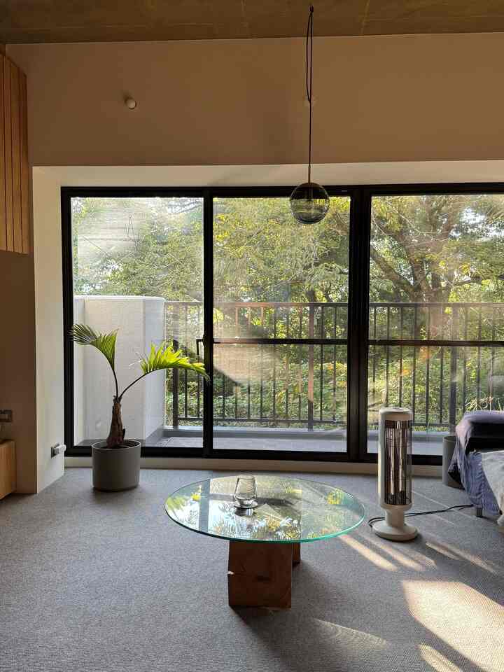 ベージュ色調のワンルームリビング、中央にガラステーブルとグレーのカーペット、暖房器具が配置された温かみのある空間