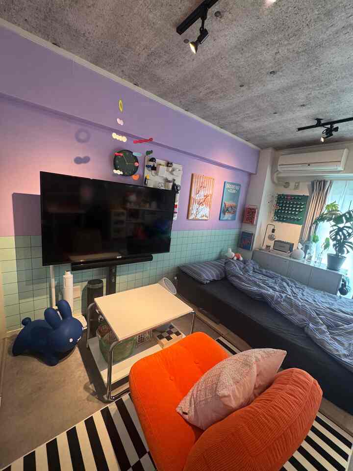 紫色と淡い緑色のトーンの小さなワンルーム、オレンジのアームチェアとベッド、テレビ、植物が配置された印象的な空間