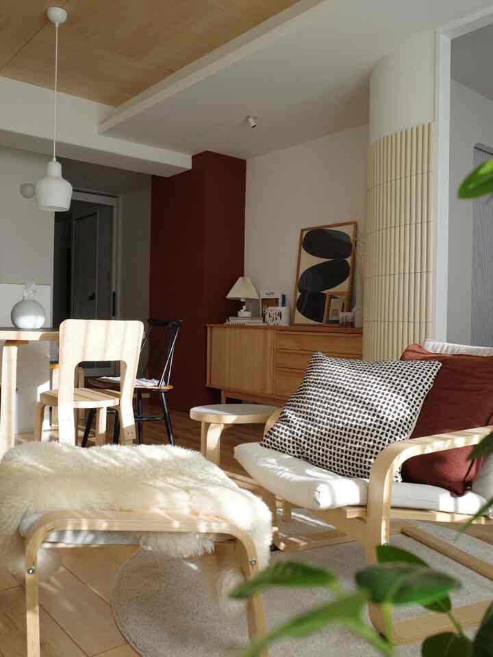 ホワイトの壁と木目調の家具が調和するリビング、オットマンとアームチェアが温かな自然光の中に配置されたナチュラルモダンなインテリア