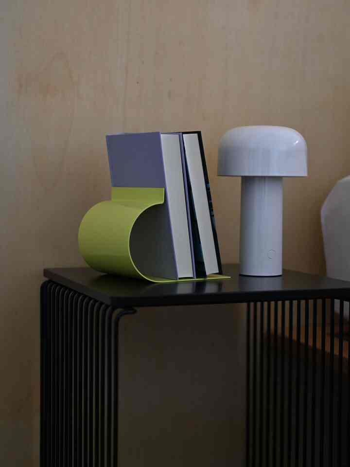 ライトグレーの壁面とブラックのメタルサイドテーブルに、グリーンとパープルのブックエンド、白いテーブルランプが配置されたモダンな寝室のインテリア