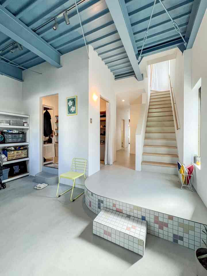 白と青の色調の玄関と階段の空間、露出したスチール棚と黄色い椅子が配置されたモダンで清潔感のあるインテリア