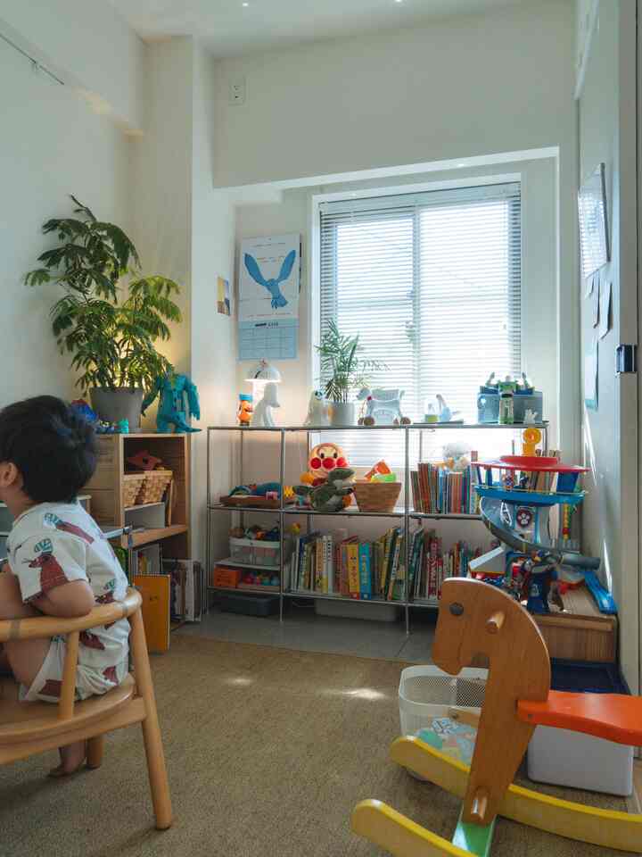 白とナチュラルカラーの子供部屋、窓際に本棚と多様なおもちゃが配置された暖かみのある空間