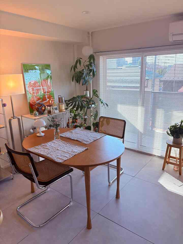 Bright white and brown-toned dining room featuring a round wooden table, rattan chairs, and plants near the window creating a cozy atmosphere