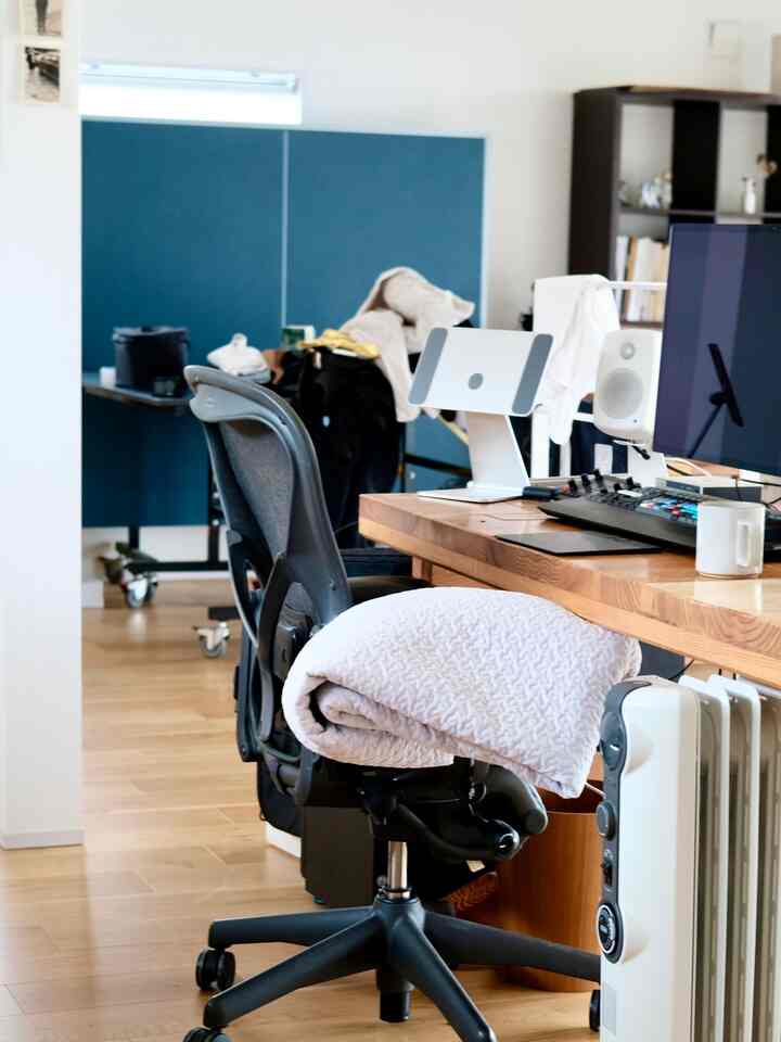 ナチュラルな木目調の床とブルーのパーティションがあるホームオフィス、ブランケットが掛けられた黒いオフィスチェアとモニタースタンドのあるデスク