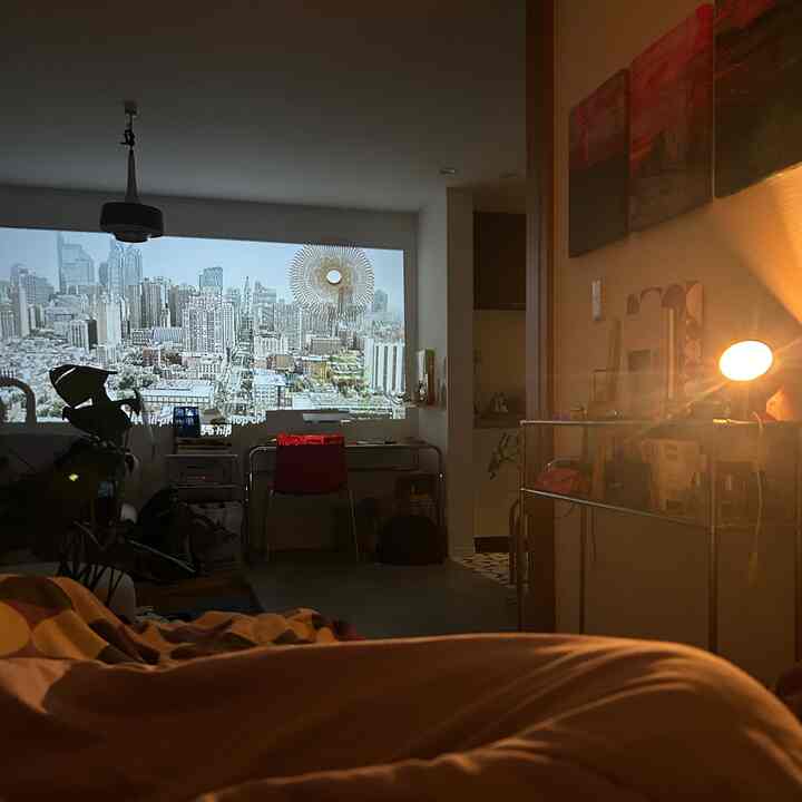 白い壁にプロジェクターが投影された1LDKの寝室兼リビングルーム、明るいフロアランプとモダンなステンレス家具が配置された落ち着いた空間