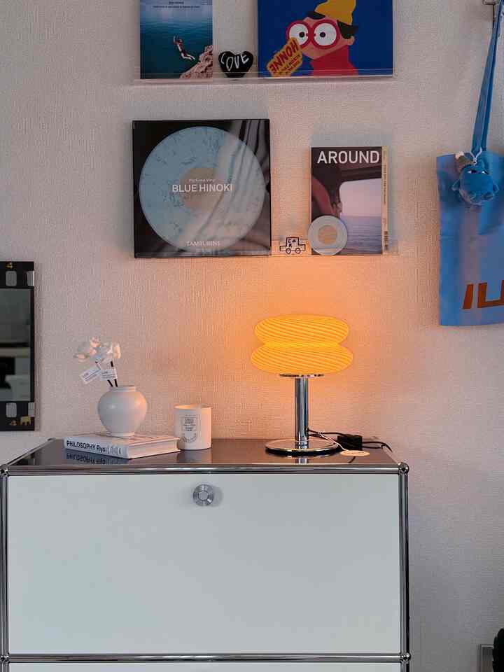 白い壁とサイドボードを背景に、黄色く輝くテーブルランプ、額縁とキャンドルが配置されたモダンな韓国インテリア