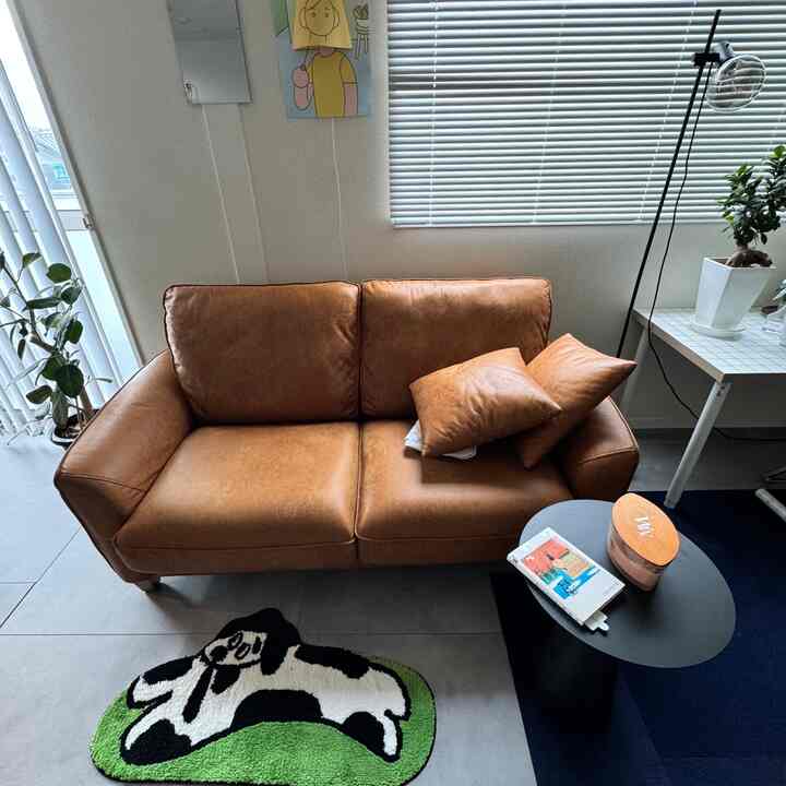 ナチュラルカラーのリビング、ブラインド付きで、ブラウンのソファと犬の形のラグ、サイドテーブルとキャンドルが配置された雰囲気のある空間