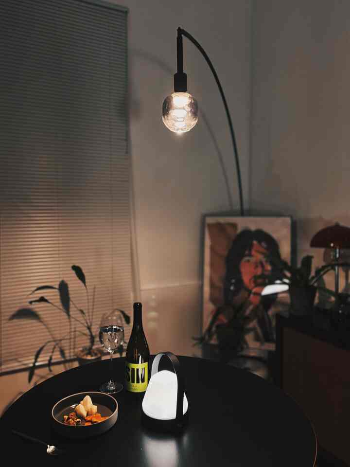 ライトグレーの壁とブラインドがあるミッドセンチュリーモダンのダイニング、中央にブラックの丸テーブルとフロアランプ、ポスターが配置されたシンプルな空間