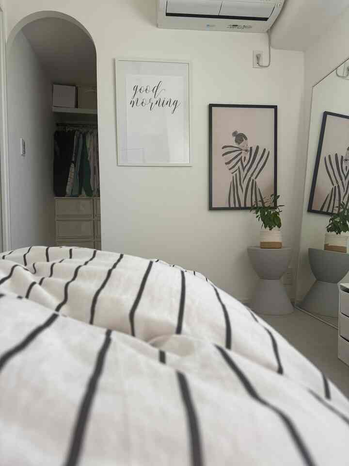 白を基調とした壁とストライプの寝具があるシンプルな寝室、額縁と収納が配置されたミニマルなインテリア