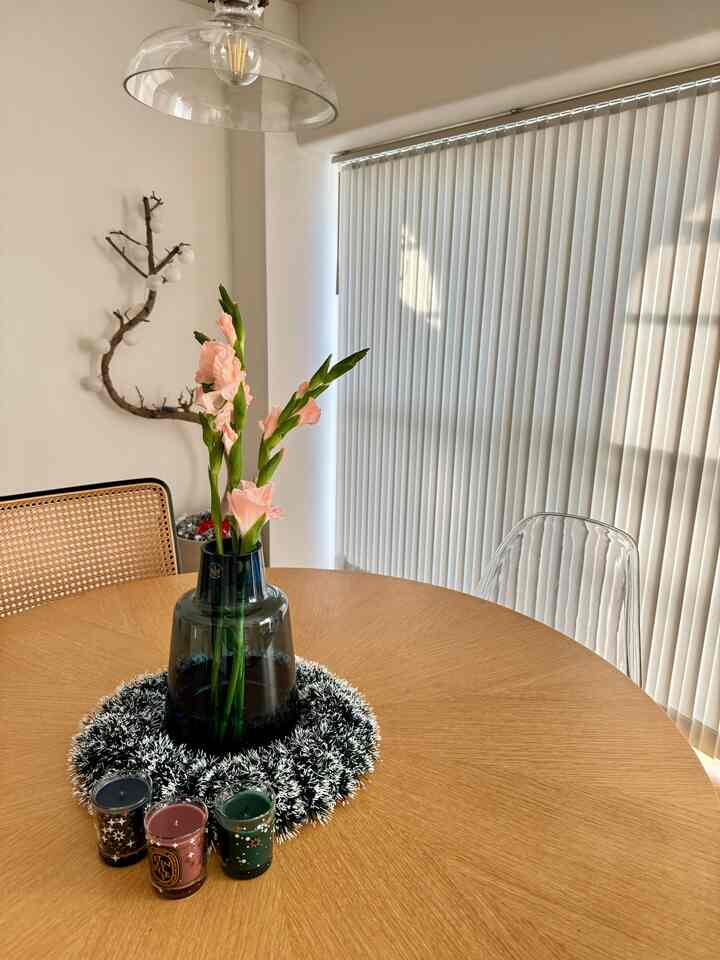 光が差し込むホワイトカーテンと木目調の丸テーブルがあるダイニングルーム、中央に花瓶とキャンドルが置かれたシンプルな空間