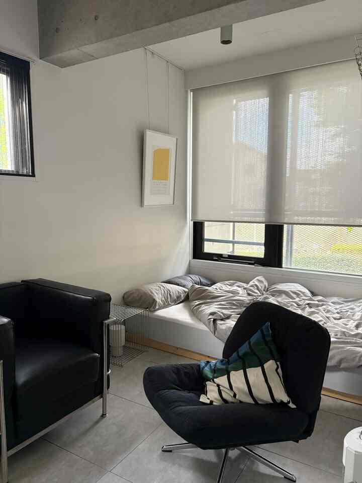 モノトーンでナチュラルモダンな寝室、黒のアームチェアとクッションのある黒いラウンジチェア、壁に黄色のアートポスターが配置された印象的な空間