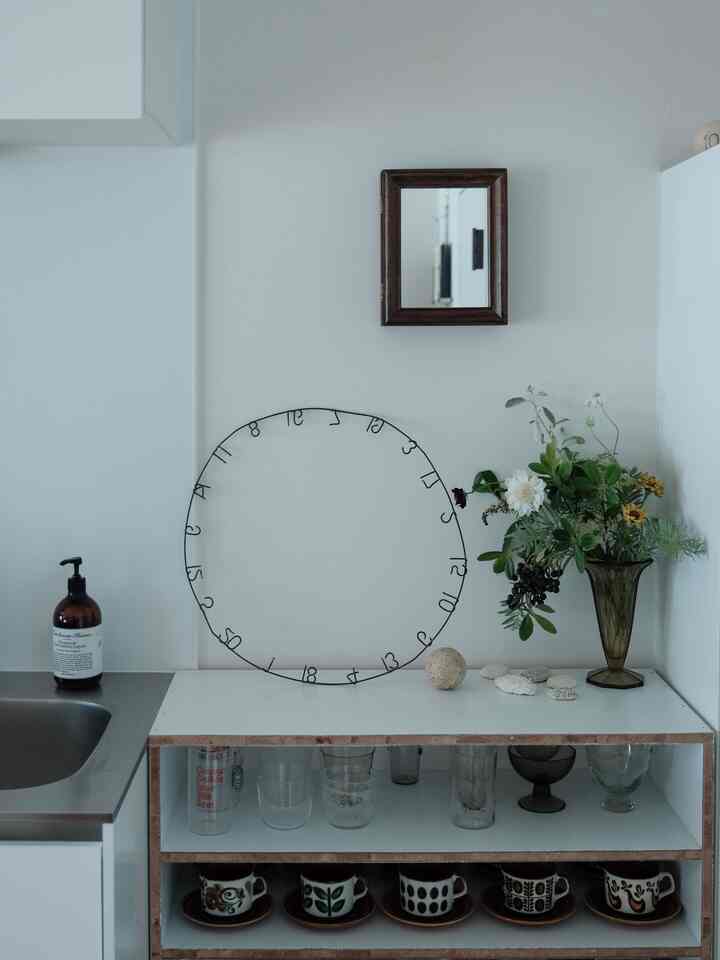 白と茶色を基調としたキッチン空間、壁中央に木製の濃い色の額縁の鏡と花瓶、開放棚に整然と並べられたカップが配置された印象的なインテリア