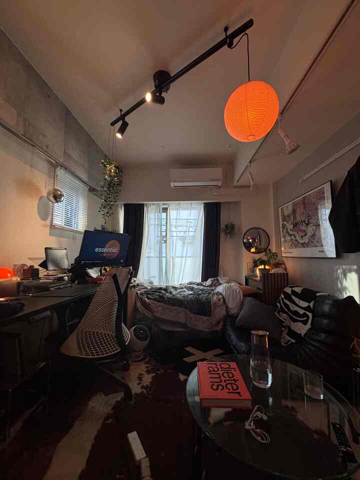 暗めのトーンの8畳ワンルーム、ガラステーブル、デスク、ベッド、ペンダントライトが配置されたモダンな一人暮らしの空間