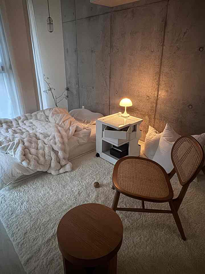A 1-person bedroom with gray concrete walls, white flooring, natural brown wooden chair and stool, and a compact white wheeled cabinet with lamp