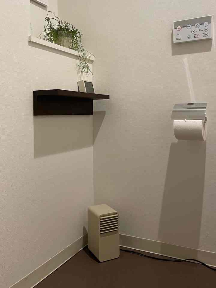 A bathroom corner with white walls and brown floor featuring a wall-mounted shelf and a compact ceramic heater in a minimal style