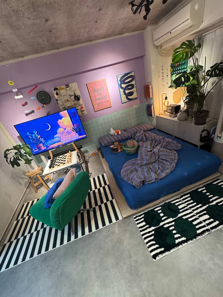 A cozy small living-bedroom space with purple walls and natural tones featuring a green armchair and indoor plants