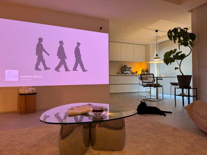 Natural beige and white living room featuring a projector screen, a black cat, and glass living table creating a cozy atmosphere