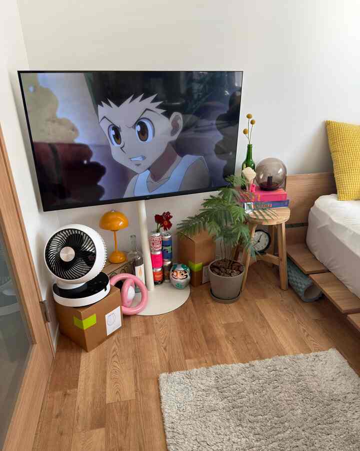 木目調の床と白い壁のあるコンパクトな一人暮らしのリビング、テレビとベッドフレーム、植物とスツールが配置された落ち着いた雰囲気のインテリア