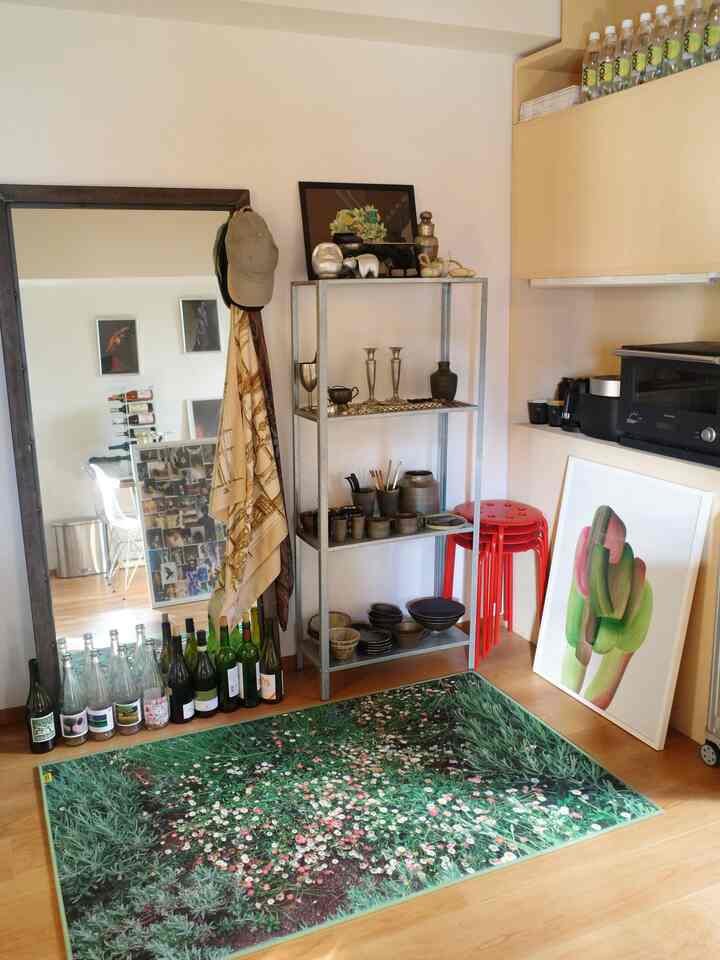 Natural-toned kitchen living area featuring a green garden rug, red stools, metal shelving with ceramics, and a large floor mirror