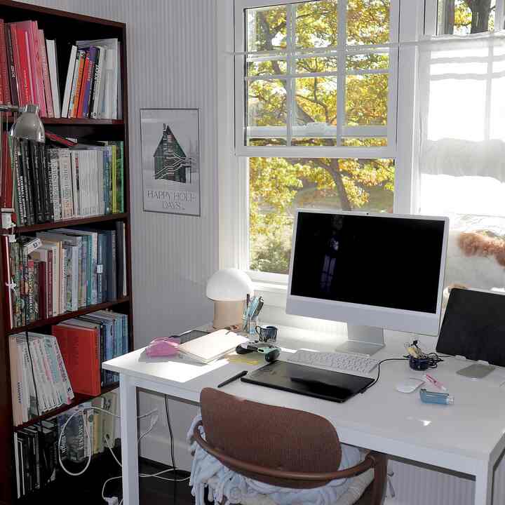 White-toned home office featuring a large window, white desk with computer, and bookshelf, creating a calm and practical atmosphere