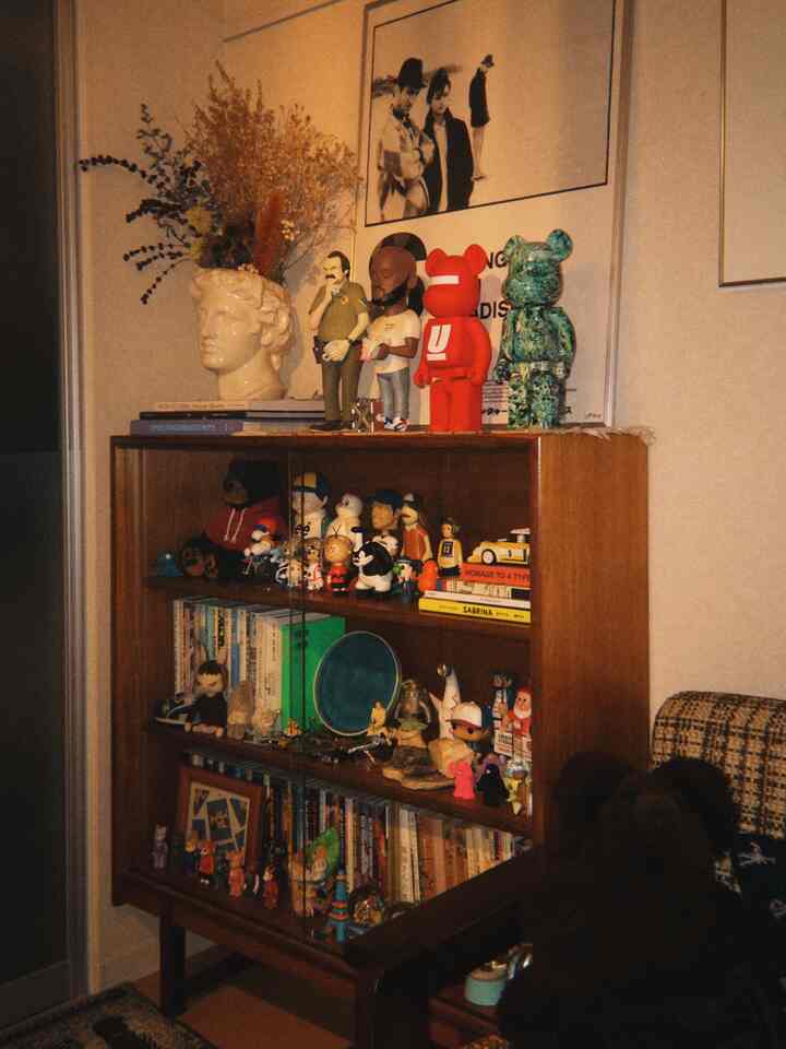 温かみのあるベージュのリビング、ヴィンテージ木製の本棚に飾られた多彩な装飾フィギュアが配置された印象的なミッドセンチュリーモダンのインテリア