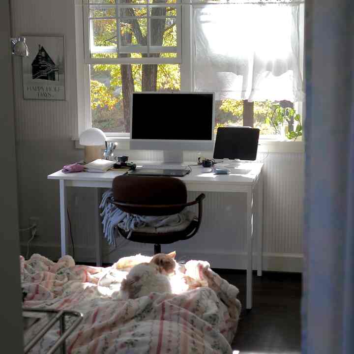 A small home office space in white and brown tones featuring a computer desk by the window with natural light and a cat resting on a cozy blanket
