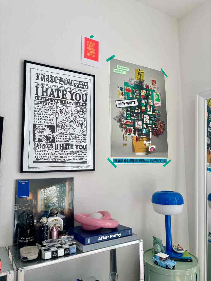 白い壁にクリスマスポスターとモノクロのアートポスターが飾られ、金属製の棚に本や小物、青いテーブルランプがある小さな一人暮らしの空間。