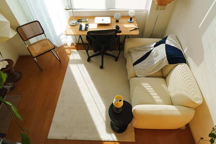 ナチュラルな木目調と白を基調とした小さなリビング、コンパクトなソファとデスクが配置された清潔感あるホームオフィス空間