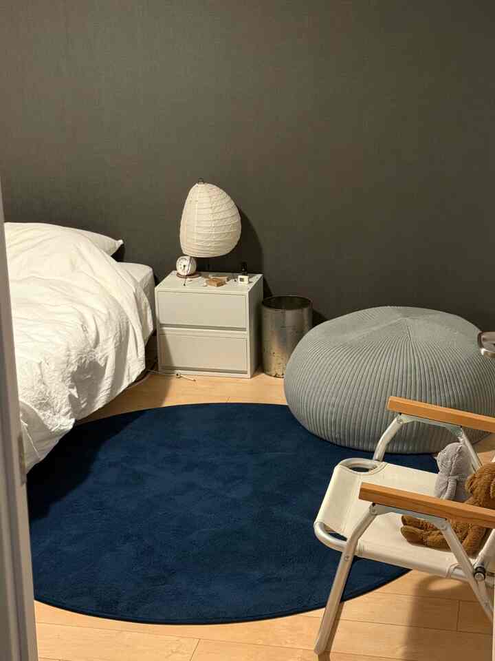 ネイビーのラグが敷かれたナチュラルな色調の寝室、白いベッドとアームチェアが配置された落ち着いた空間