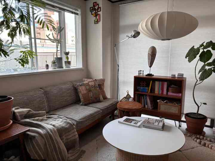 ベージュとブラウンの色調が調和したリビングルーム、木製の本棚とソファ、ペンダントライト、そして多くの観葉植物が配置された雰囲気のあるインテリア