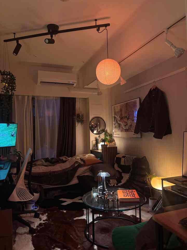 暖かいオレンジ色の間接照明が照らす8畳のモダンなワンルーム、ベッドやソファ、丸いガラスのコーヒーテーブルが配された雰囲気のある空間