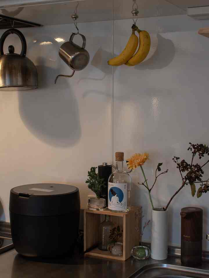 白いタイルの壁と黒い炊飯器がある整ったキッチン、花瓶や吊るされたバナナが飾られている雰囲気のある空間