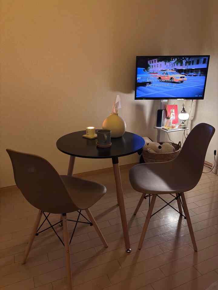 ナチュラルカラーとブラックの調和が取れた二人用ダイニングルーム、丸テーブルと椅子、壁掛けテレビが配置された落ち着いた空間