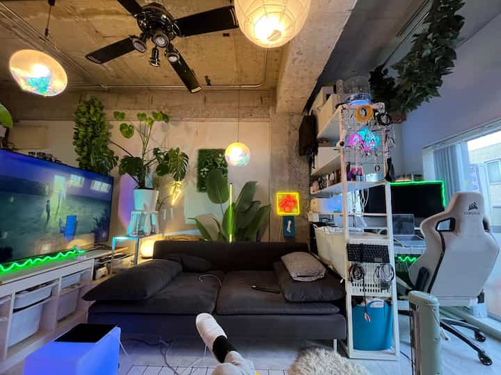 黒いソファと白いゲーミングチェアが配置されたワンルームのリビング、多数の観葉植物とペンダントライトが温かみのある雰囲気を演出する居心地の良い空間