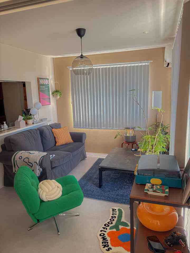 ベージュの壁とライトグレーの床が特徴的な8畳のリビング、グレーのソファとグリーンのアームチェア、コーヒーテーブルと植物が配置された雰囲気のある空間