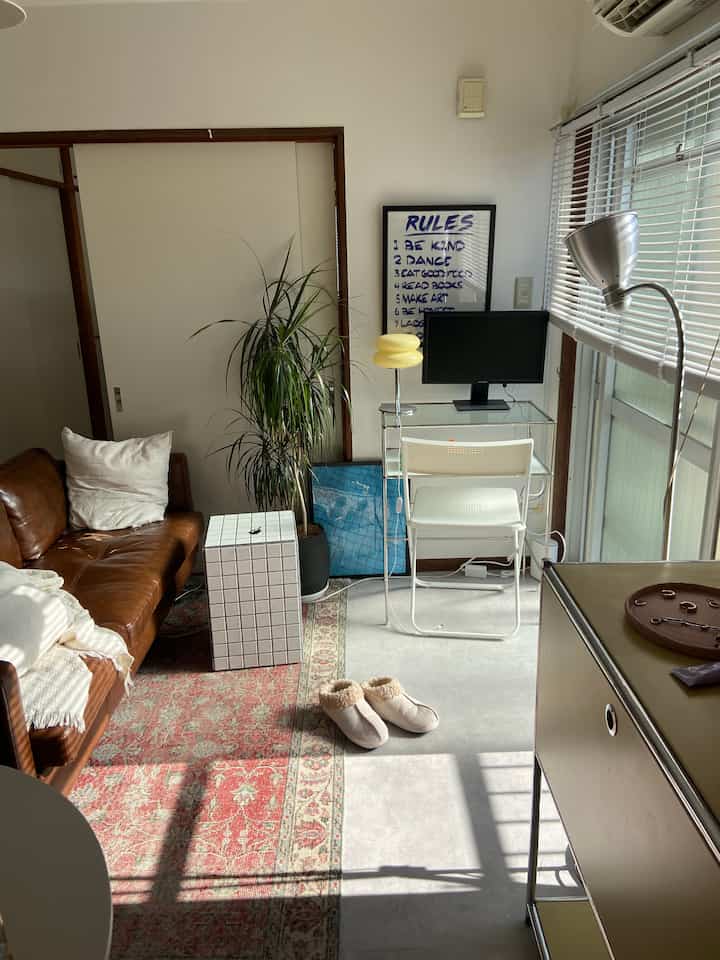 ナチュラルカラーの一人暮らしリビング、ブラウンのソファとホワイトのブラインド、ワークデスクが配置された温かみのあるインテリア