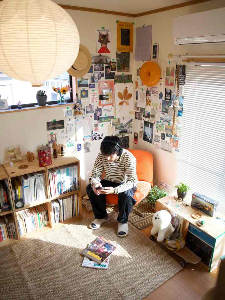 ナチュラルなトーンの6畳リビング、オレンジのアームチェアとラグ、多彩なポスターと植物が配置された雰囲気のある空間