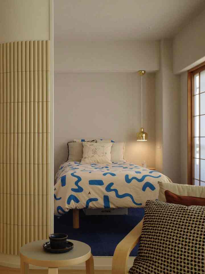 Light brown and navy toned bedroom featuring Artek bed and stool with golden pendant lamp in a simple setting