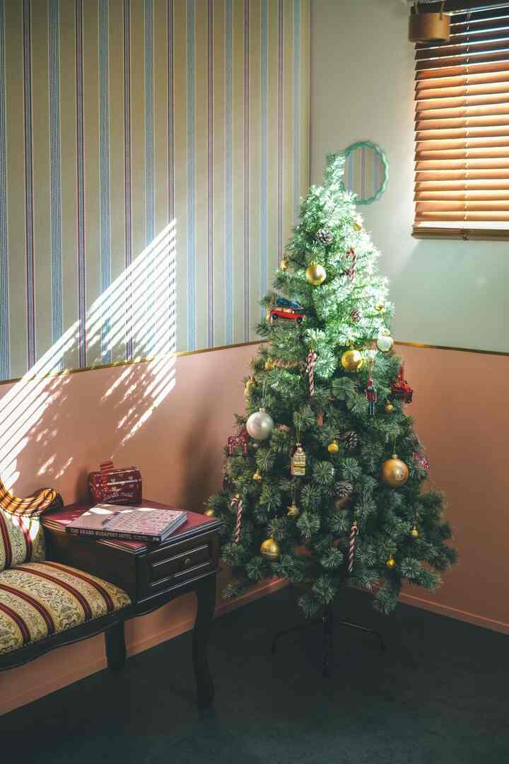 A living room with soft pink walls and striped wallpaper features a Christmas tree decorated with gold and red ornaments under warm sunlight.