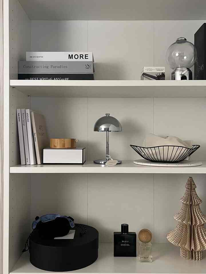 白い壁と本棚を背景に、クロームのテーブルランプと木製オブジェが配置されたミニマルなインテリアの棚