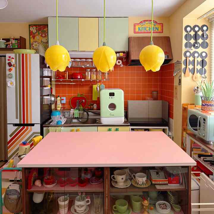 黄色のペンダントライトとオレンジのタイル壁が印象的なレトロなキッチン、ピンクのダイニングテーブルと多彩なキッチン用品が配置された温かみのある空間